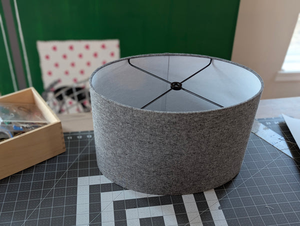 oval lamp shade in grey fabric with black hardware sits on a work table on a cutting mat with tools and supplies around it