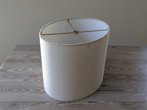 oval shaped lampshade in a speckled natural fabric with brass hardware sitting on a light wood table