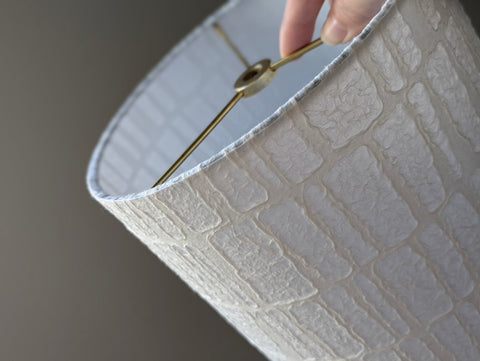 Close-up of a textured paper lampshade with a repeating trapezoid pattern and a hand holding it by the brass hardware