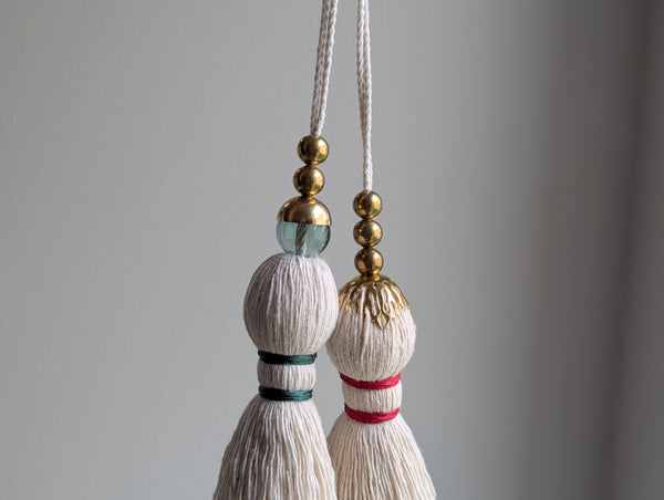 A decorative tassel featuring off-white cotton cord, two small metallic gold beads, one larger metallic gold bead, and a translucent green glass bead