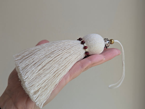 A hand holding a decorative cream-colored tassel door hanger with small dark red beads and a gold accent bead, against a plain beige background.