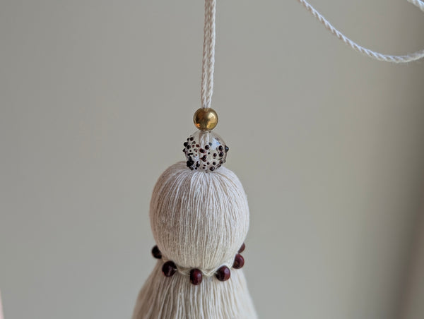 A close-up of a decorative hanging ornament with a cream cotton tassel, a small gold bead, a burgundy spotted bead, suspended by a white cord against a soft beige background.