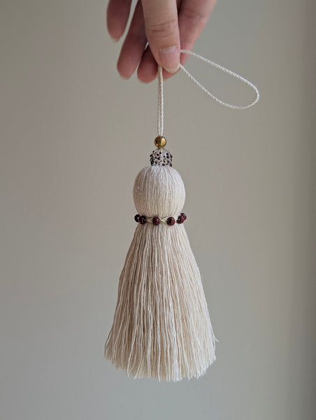 A hand holding a decorative hanging ornament with a cream-colored tassel, dark red beads, and a small gold bead against a plain light gray background.