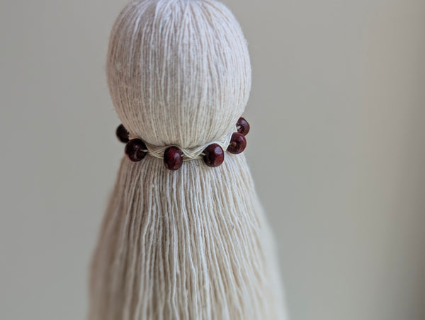 A close-up of a cream-colored cotton tassel wrapped with a dark red beaded band, set against a soft, neutral beige background.
