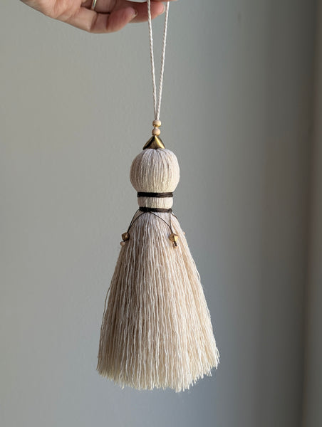 A close-up of a decorative tassel with cream-colored thread, a brown wrapped detail, and brass and wooden beads shown against a neutral gray background