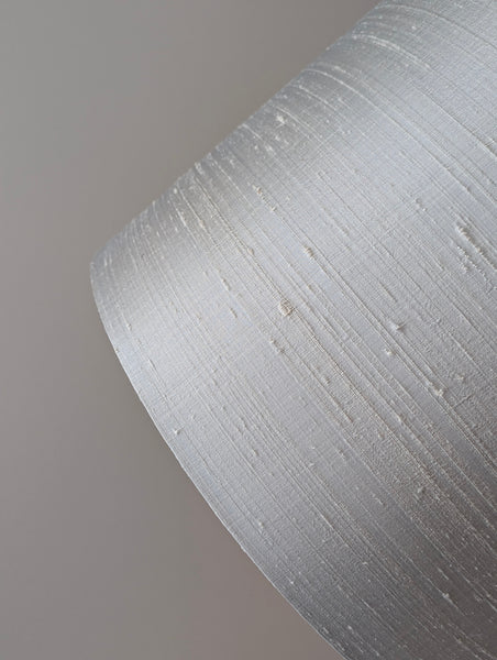 A close-up of a textured gray silk lamp shade with a fine horizontal nubby pattern, set against a solid neutral gray background.