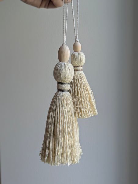 A close-up of a decorative tassel with cream-colored thread, a tan wrapped band, and a wooden beads shown against a neutral gray background