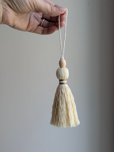 A close-up of a decorative tassel with cream-colored thread, a tan wrapped band, and a wooden beads shown against a neutral gray background