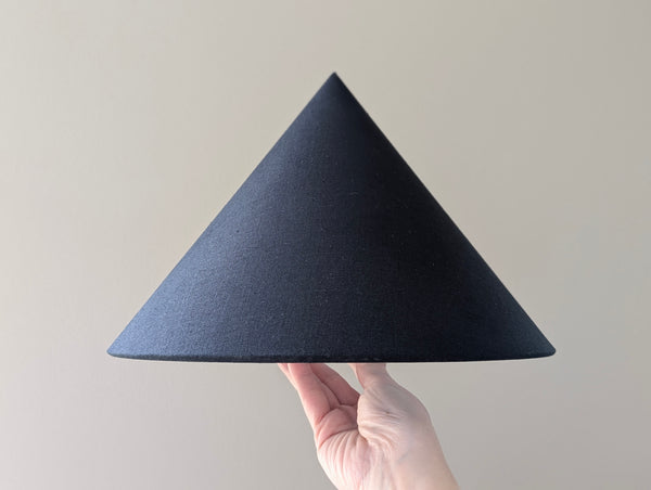 A hand holding a black linen cone lamp shade against a plain light beige background.