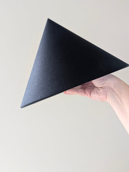 A hand holding a black linen cone lamp shade against a plain light beige background.