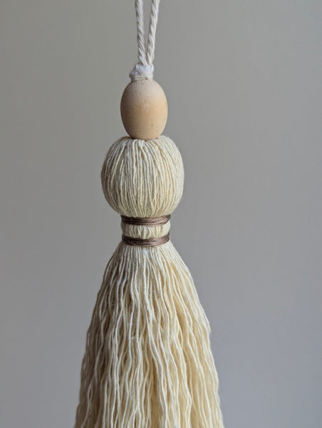 A close-up of a decorative tassel with cream-colored thread, a tan wrapped band, and a wooden beads shown against a neutral gray background