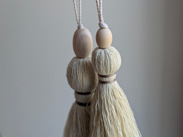 A close-up of a decorative tassel with cream-colored thread, a tan wrapped band, and a wooden beads shown against a neutral gray background