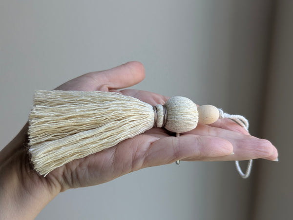 A close-up of a decorative tassel with cream-colored thread, a tan wrapped band, and a wooden beads shown against a neutral gray background