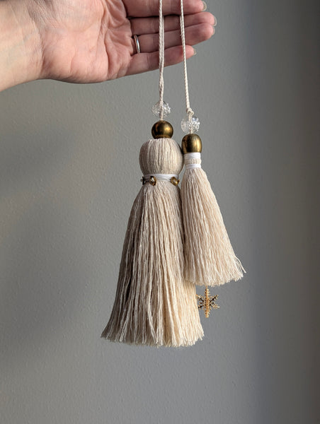 snowy white snowflake tassel with white speckled glass bead and brass snowflake charm hanging next to a larger tassel with brass and glass accents