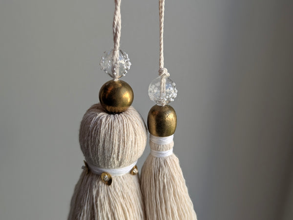 Two cream-colored cotton tassels hanging from white cords with clear and white speckled beads and metallic gold accents, set against a neutral gray background.