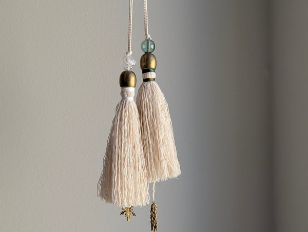 Two cream-colored cotton tassels hanging from white cords with brass and glass bead accents against a neutral gray wall.