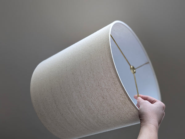 Speckled Oatmeal cotton fabric lampshade with brass hardware against a blurred background