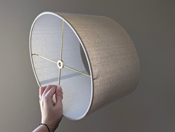 hand holding oval shaped lamp shade with brass hardware and natural colored cotton fabric