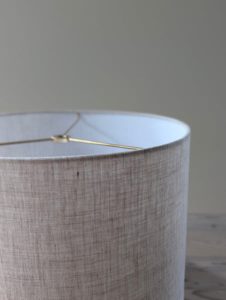 Close-up of a woven linen lamp shade with brass hardware against a blurred background