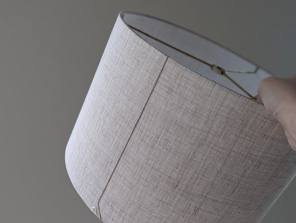 the back seam of a tan linen lamp shade with brass hardware being held by a hand