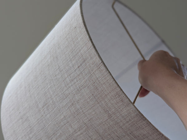 Close-up of a light tan linen lampshade with brass hardware being held by a hand