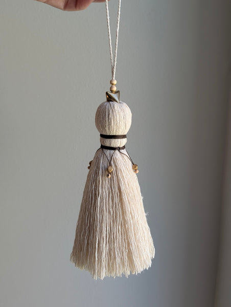 A close-up of a decorative tassel with cream-colored thread, a brown wrapped detail, and brass and wooden beads shown against a neutral gray background
