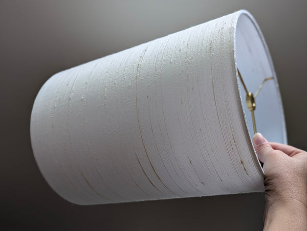 A hand holding a white drum fabric lampshade brass hardware, shown against a dark gray background.