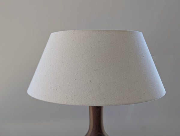 A close-up of a table lamp shade made from textured, speckled off-white fabric, mounted on a walnut lamp against a neutral gray background.