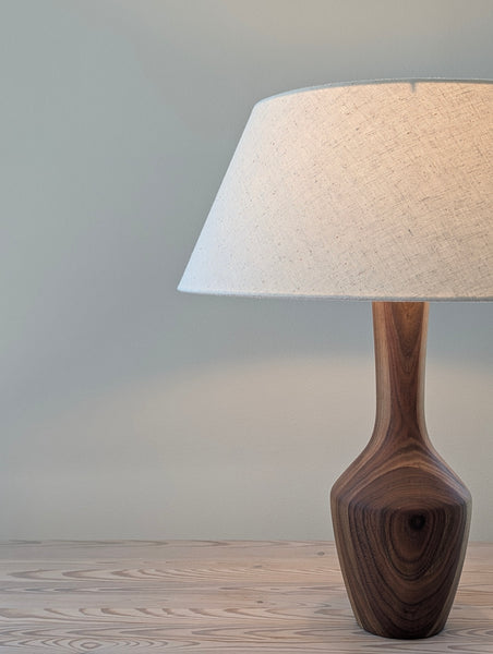 A table lamp with a dark walnut wood base and a textured cream fabric drum shade, shown on a light wood surface against a neutral gray wall.