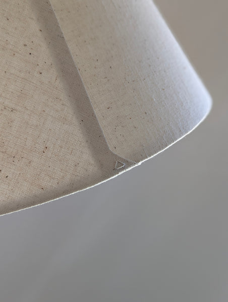 Close-up of a textured off-white fabric lampshade with subtle brown speckles, showing the curved edge and fine weave against a neutral background.