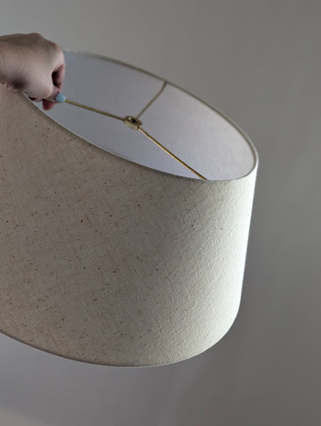 A close-up of a large drum-shaped lampshade with a textured off-white linen fabric, held by a hand at the top edge