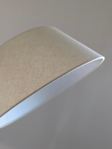 A close-up of a curved lampshade made of light beige linen fabric, showing its textured weave and white inner lining against a neutral gray background.
