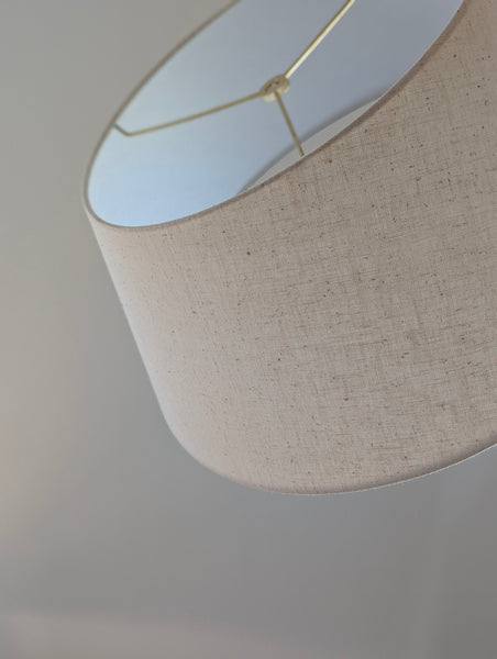 Close-up of a drum lampshade made of textured beige linen fabric, with brass spider washer hardware visible inside, set against a soft neutral gray background.