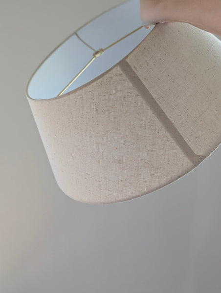 A close-up of a drum-shaped fabric lampshade in a light beige linen-like material, with a brass spider washer visible inside the top opening against a plain light gray background.