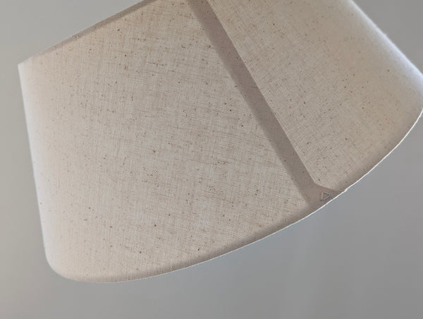 A close-up of a tapered fabric lampshade made of a textured, speckled beige linen-like material, showing the seam and edge details against a neutral gray background.