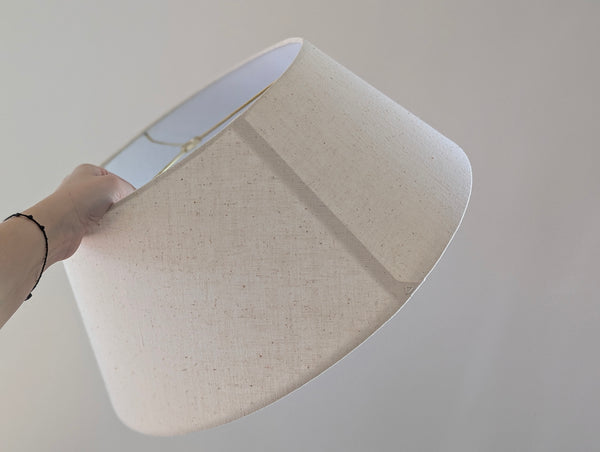 A hand holding a large drum-shaped fabric lampshade with a natural linen texture, set against a plain light gray background.