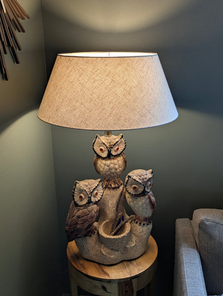 A decorative table lamp shaped like three owls perched on a branch, with a beige textured drum shade, shown lit in a real interior setting beside a chair.