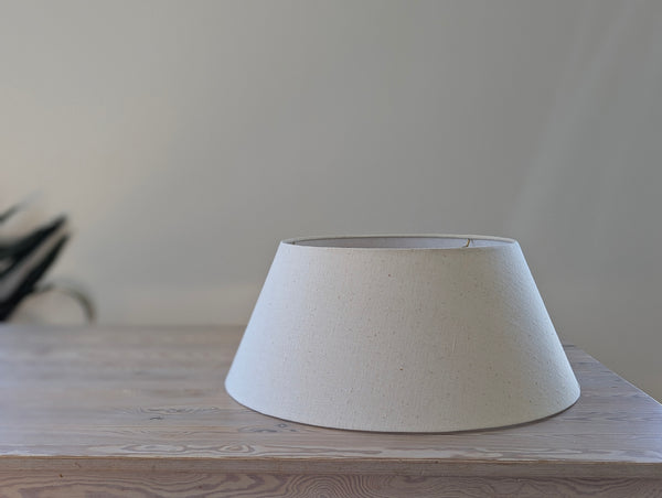 A natural beige linen lampshade with a tapered conical shape, displayed on a light wood tabletop against a neutral, softly blurred background.