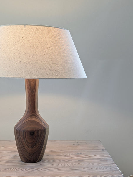 A table lamp with a simple walnut base and a tapered beige linen shade, shown lit on a light wood tabletop against a soft gray wall.