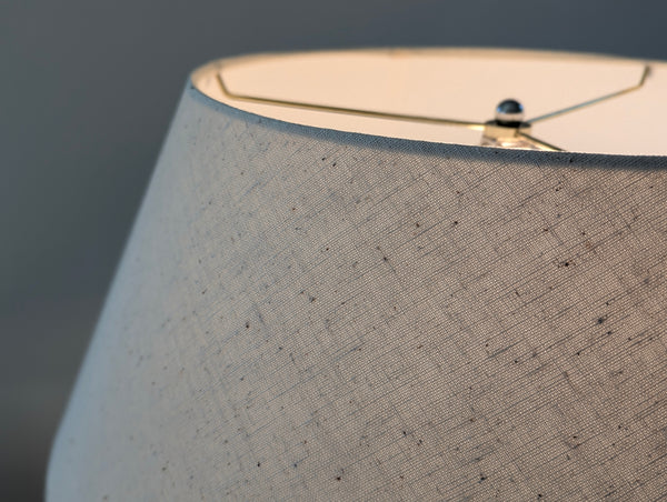 Close-up of a textured fabric lampshade in a warm light beige or off-white tone, showing a fine woven pattern with small dark flecks and a metal top finial.