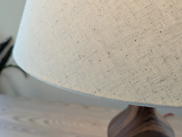 Close-up of a textured off-white linen lampshade with small speckles, showing the fabric weave and edge, with a dark wooden lamp base partially visible below on a light wood surface.