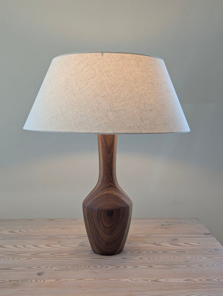 A table lamp with a tapered off-white linen shade and a sculptural dark walnut wood base, shown lit on a light wood tabletop against a neutral gray wall.