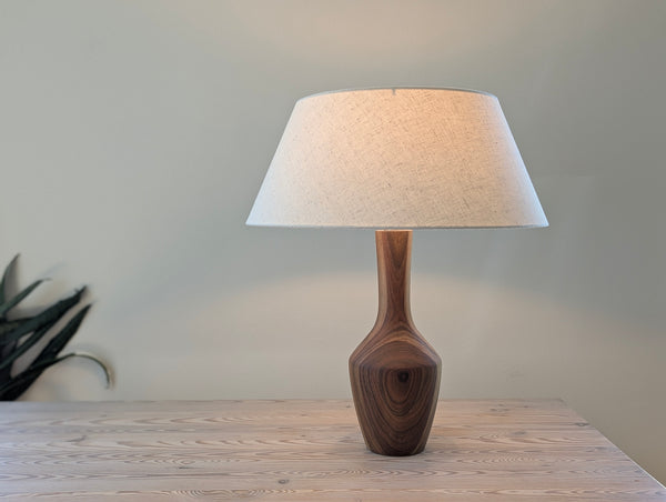 A table lamp with a textured beige fabric drum shade and a sculptural walnut wooden base, shown lit on a light wood tabletop against a neutral wall.