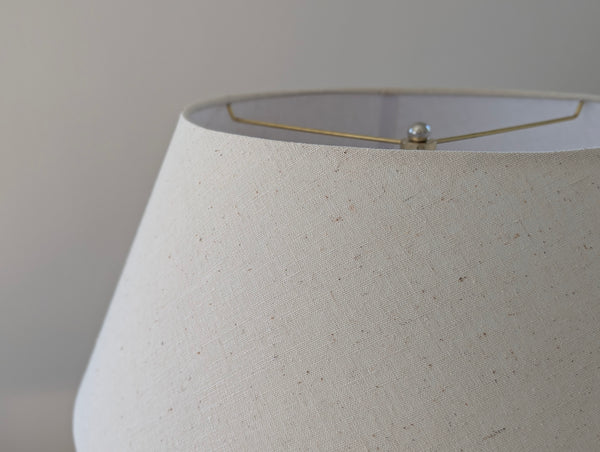 Close-up of a tapered fabric lampshade in a warm off-white or cream color with subtle speckles, showing a brass spider washer frame and a small finial at the top rim.