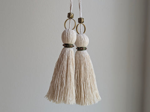 Two cream-colored cotton tassels with brass rings and decorative bands, hanging against a plain light gray wall.