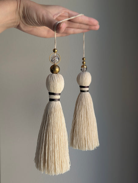 A close-up of a double tassel with deep purple colored thread, and glass and brass beads at the top against a neutral gray background