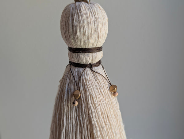 A close-up of a decorative tassel with cream-colored thread, a brown wrapped detail, and brass and wooden beads shown against a neutral gray background