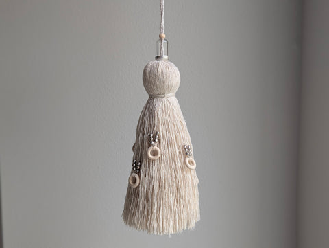 A close-up of a decorative tassel with cream-colored thread, a silver beads with wooden loops hanging from the middle, and glass, silver and wooden beads at the top shown against a neutral gray background