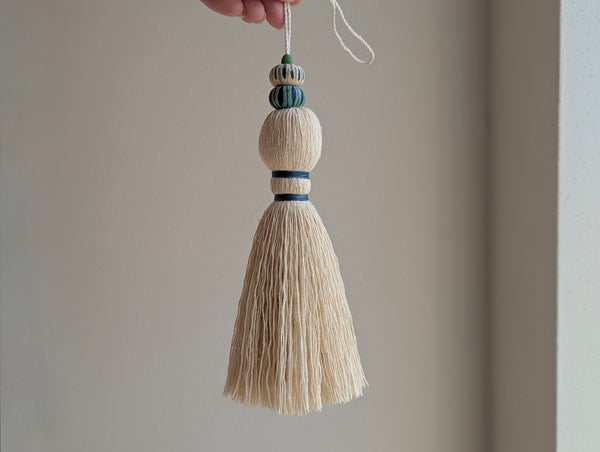 A close-up of a handmade wooden bead tassel, featuring a cream-colored cotton and blue and green accents against a soft beige background.