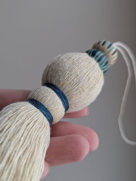 A close-up of a handmade wooden bead tassel, featuring a cream-colored cotton and blue and green accents against a soft beige background.